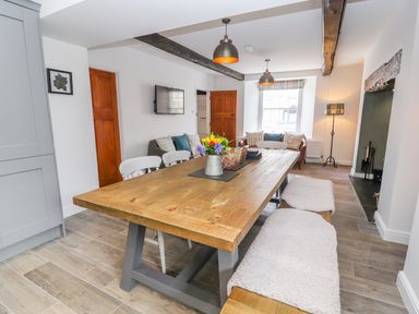 A dining room with a wooden table and benches with cushions a sofa a mounted tv and a window at Llys Alaw in Llangernyw