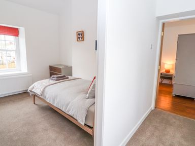 A bedroom with a single bed with folded towels on top a chest of drawers a window with a red blind and a view through the door to another room with a lamp and wardrobe at Llys Alaw in Llangernyw