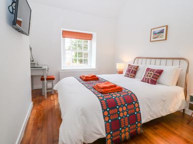 A bedroom with a bed with white bedding and patterned runner and pillows a window with an orange blind a small desk with a stool and a wall-mounted television at Llys Alaw in Llangernyw