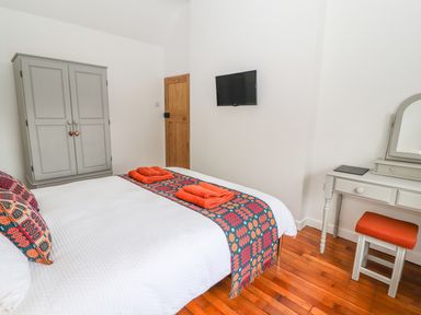 A bedroom with a bed covered in white bedding and colorful blankets a gray wardrobe a wooden door a wall-mounted TV and a white dressing table with an orange stool at Llys Alaw in Llangernyw