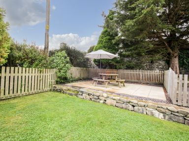 A fenced garden with a grassy lawn stone wall patio table with umbrella and trees at Llys Alaw in Llangernyw