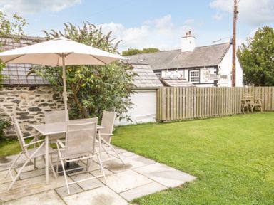 An outdoor patio with a table and six chairs under an umbrella next to a stone wall and green lawn at Llys Alaw in Llangernyw