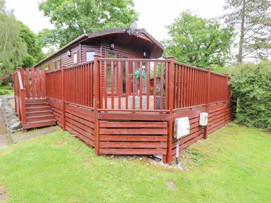 A wooden cabin with a large deck and railing in a grassy area with trees at 10 Thirlmere White Cross Bay near Troutbeck Bridge