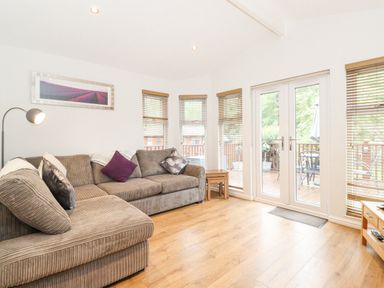 A living room with a brown sectional sofa and pillows beside windows and glass doors leading to a deck at 10 Thirlmere White Cross Bay near Troutbeck Bridge