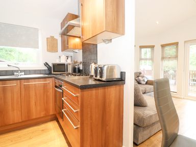 A kitchen with wooden cabinets a stove microwave and toaster next to a living area with a sofa and chair at 10 Thirlmere White Cross Bay near Troutbeck Bridge