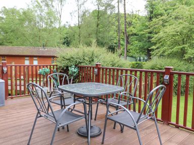 An outdoor wooden deck with a square glass table and four metal chairs surrounded by red railing and green trees at 10 Thirlmere White Cross Bay near Troutbeck Bridge