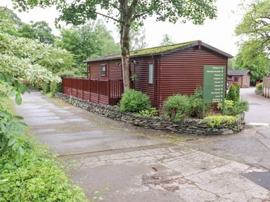 A wooden cabin with a mossy roof next to a stone wall and green directional sign surrounded by trees and plants at 10 Thirlmere White Cross Bay near Troutbeck Bridge