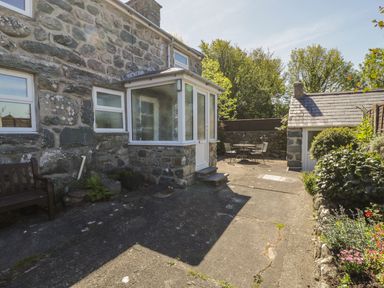 An outdoor stone patio area with a bench a small porch door a garden shed table and chairs and various plants at Ty Uchaf in Dyffryn Ardudwy