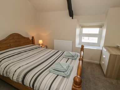 A bedroom with a wooden bed, striped bedding, folded towels, a small bedside lamp, a window, and a chest of drawers at Ty Uchaf in Dyffryn Ardudwy