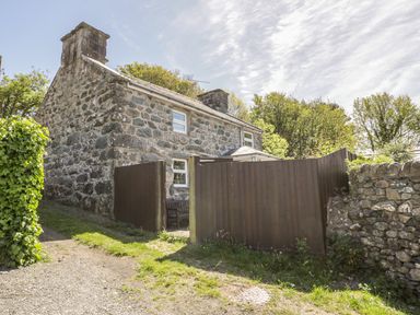 A stone house with a chimney and wooden fence surrounded by trees and grass at Ty Uchaf in Dyffryn Ardudwy