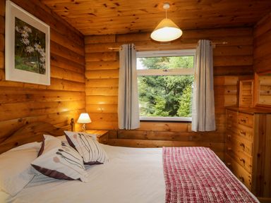A bedroom with a wooden bed white and patterned pillows a nightstand with a lamp a window with curtains and a wooden dresser with mirrors at Eagle Lodge in Strathpeffer