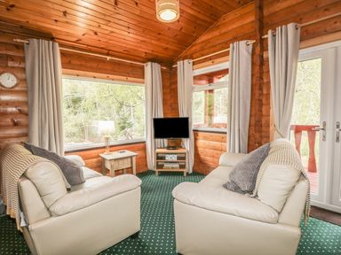 A living room with two cream sofas a TV on a stand a side table with a lamp and large windows at Otter Lodge in Strathpeffer