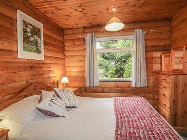 A bedroom with a wooden bed frame a bedside table with lamp a chest of drawers and a window with curtains at Otter Lodge in Strathpeffer
