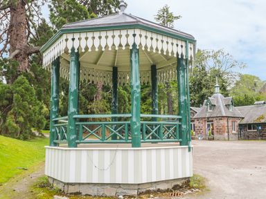 A green and white gazebo with decorative trim in a garden with trees and stone buildings in the background at Otter Lodge in Strathpeffer