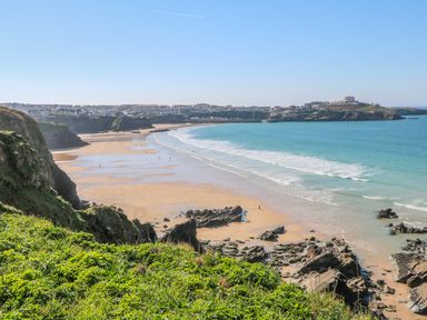 A sandy beach with people walking near the shore and rocky cliffs with green grass overlooking the ocean at Ocean Seven in Newquay