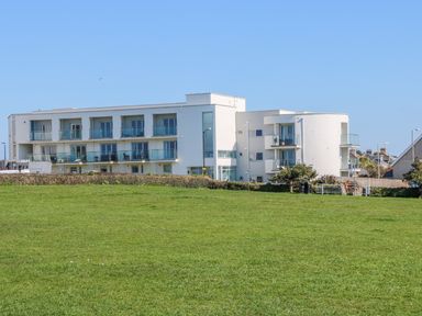 A modern white building with balconies and large windows behind a green grassy field at Ocean Seven in Newquay