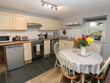 A kitchen with cream cabinets a round dining table with flowers on it and kitchen appliances at Bwthyn Y Jacdo in Crymych