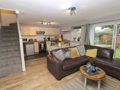 An open living room and kitchen with a brown leather sectional sofa cushions a wooden coffee table with wine glasses and a bottle a dining table with chairs flowers on it white cabinets kitchen appliances and carpeted stairs visible at Bwthyn Y Jacdo in Crymych