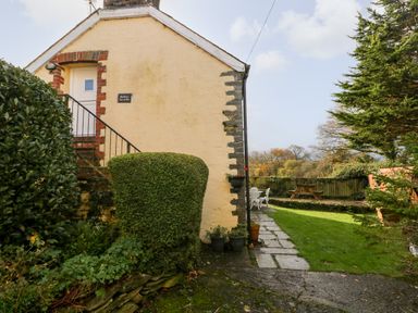 The exterior of a house with a small staircase and door surrounded by bushes and a garden with a path and outdoor furniture at Bwthyn Y Jacdo in Crymych
