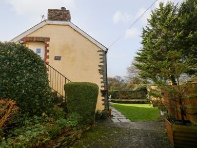 The side of a yellow brick house with stairs and green bushes beside a garden with a tree picnic table and a small lawn at Bwthyn Y Jacdo in Crymych