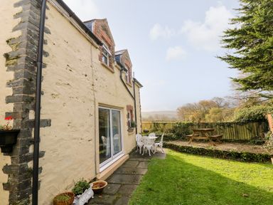 A garden area with a yellow stone house patio metal table and chairs and wooden picnic bench surrounded by grass and trees at Bwthyn Y Jacdo in Crymych