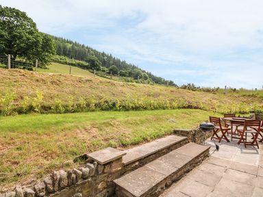 A patio with wooden table and chairs and a barbecue grill overlooking a grassy hill and trees at The Barn in Abergavenny