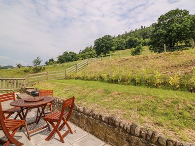 An outdoor patio with a wooden table and chairs next to a grassy hill and wooden fence at The Barn in Abergavenny