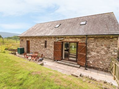 An outdoor patio with a round wooden table and chairs next to a stone barn with wooden doors at The Barn in Abergavenny