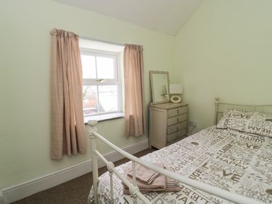 A bedroom with a bed linens printed with words a small chest of drawers with a lamp and mirror and a window with curtains at Preswylfa in Penmachno