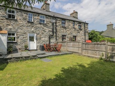 A stone house with a small patio and wooden chairs and table in a fenced garden at Preswylfa Penmachno