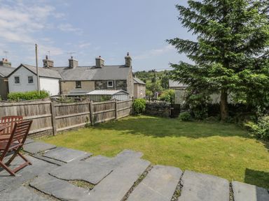 A garden with a lawn wooden fence stone patio table and chairs large tree and houses in the background at Preswylfa in Penmachno