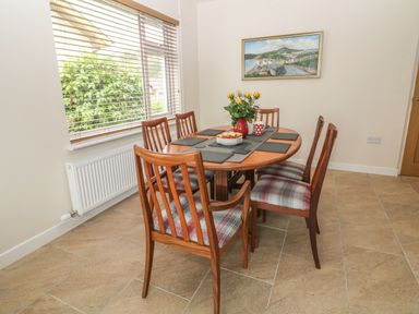 A dining area with a wooden table and six chairs with plaid cushions by a window with blinds and a painting on the wall at Cae Glas in Pwllheli