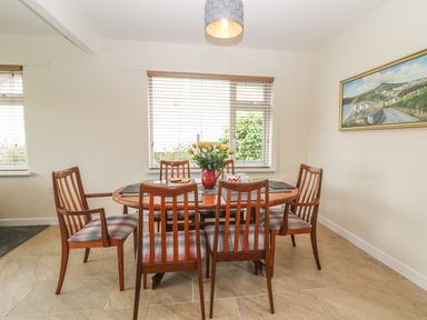 A dining room with a wooden oval table and six chairs with plaid cushions yellow flowers in a red vase on the table and a landscape painting on the wall at Cae Glas in Pwllheli