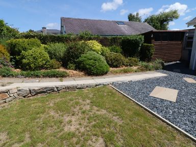 A garden with grass a stone wall bushes a wooden shed and a gravel area with stepping stones at Cae Glas in Pwllheli