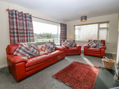A living room with red leather sofa and chairs with plaid cushions a red rug on a grey carpet and large windows with blinds and curtains at Cae Glas in Pwllheli