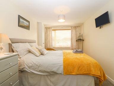 A bedroom with a bed white sheets yellow blanket pillows a bedside table lamp a wall mounted TV and window blinds at Cae Glas Pwllheli