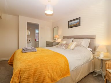 A bedroom with a large bed with yellow blanket and pillows bedside tables with lamps a chest of drawers and an open door to a bathroom at Cae Glas in Pwllheli
