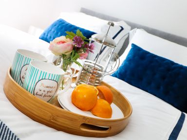 A wooden tray with mugs, a French press, flowers, and oranges on a bed with blue cushions at An Drowes in Newquay