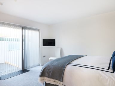A bedroom with a bed covered with a blanket a white chair a wall-mounted TV and vertical blinds on sliding glass doors at An Drowes in Newquay