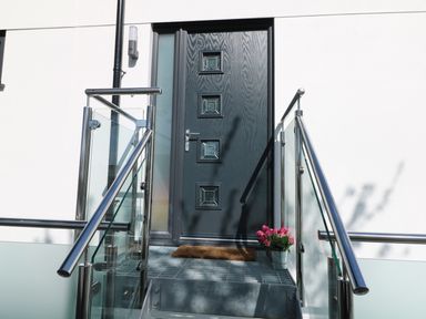 A black front door with glass panes and a metal railing with glass panels on stairs and a potted plant beside the door at An Drowes in Newquay