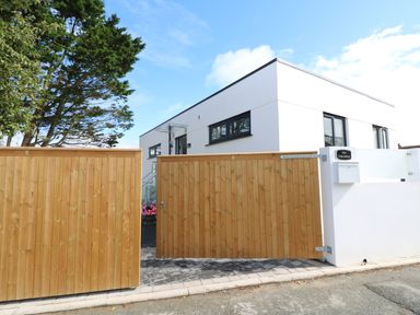 A front gate made of wood with a modern white building behind it at An Drowes in Newquay