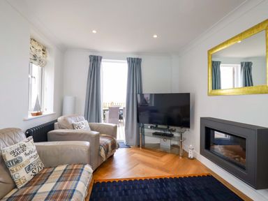 A living room with two armchairs a television on a glass stand a wall-mounted fireplace and a large mirror at Pink House in Mevagissey