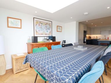 A dining room with a long table covered by a patterned tablecloth surrounded by chairs and a wooden sideboard with framed maps on the wall at Pink House in Mevagissey