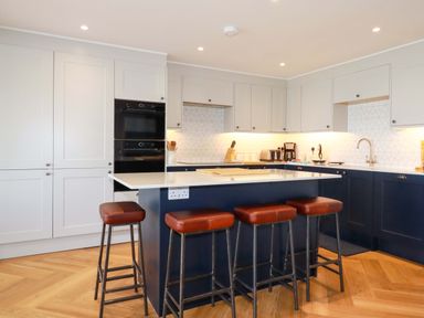 A kitchen with white and dark blue cabinets a white countertop an island with four brown stools and wooden flooring at Pink House in Mevagissey