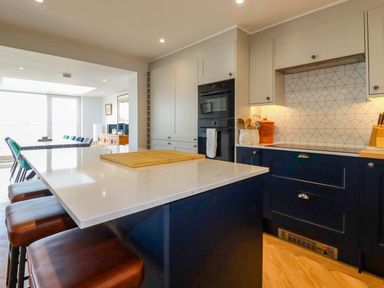 A kitchen with a white countertop island and brown stools next to blue and white cabinets with built in ovens and a cutting board at Pink House in Mevagissey