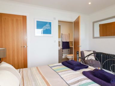 A bedroom with a bed, towels on the bed, two wooden doors, a chair with a cushion, a wall mirror and an en suite bathroom at Pink House Mevagissey