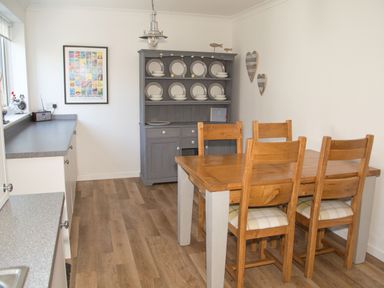 A dining area with a wooden table and four chairs next to a grey cabinet with plates and bowls on display at Seashell Cottage in Amble-By-The-Sea