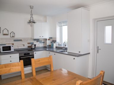 A kitchen with white cabinets wooden chairs and a wooden table at Seashell Cottage in Amble-By-The-Sea