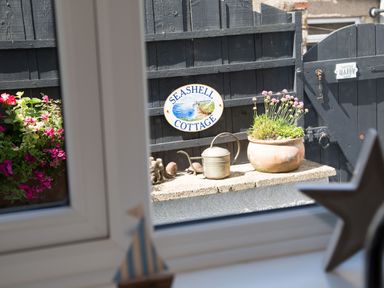 A garden area with a flower pot watering can and a seashell cottage sign viewed through a window at Seashell Cottage in Amble-By-The-Sea