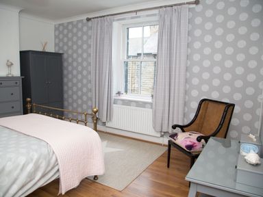 A bedroom with a brass bed frame gray wardrobe a wooden chair with a cushion and a window with curtains at Seashell Cottage in Amble-By-The-Sea